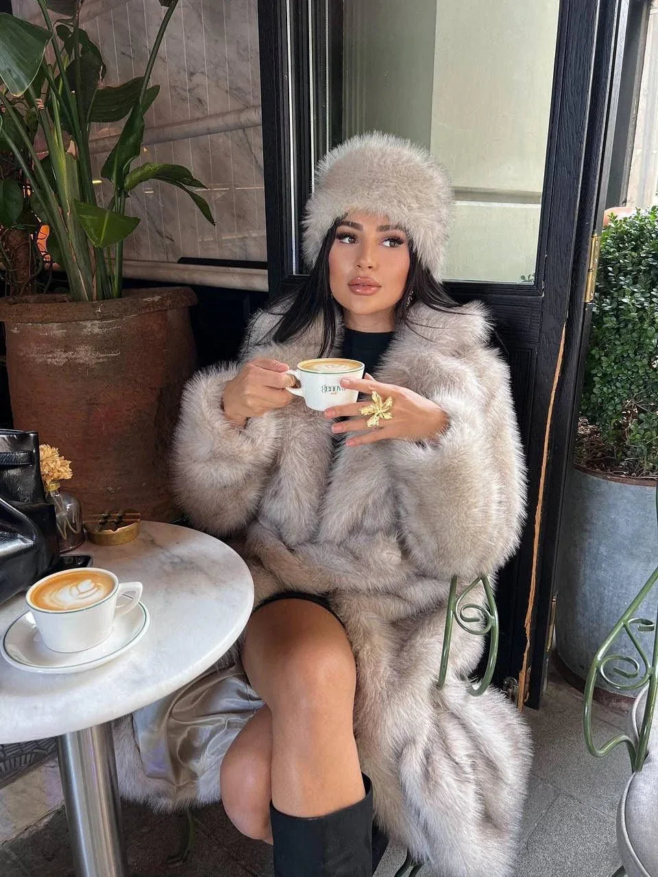 Woman wearing a long, cream-colored faux fur coat and a matching faux fur hat, sitting at a cafe table with coffee.