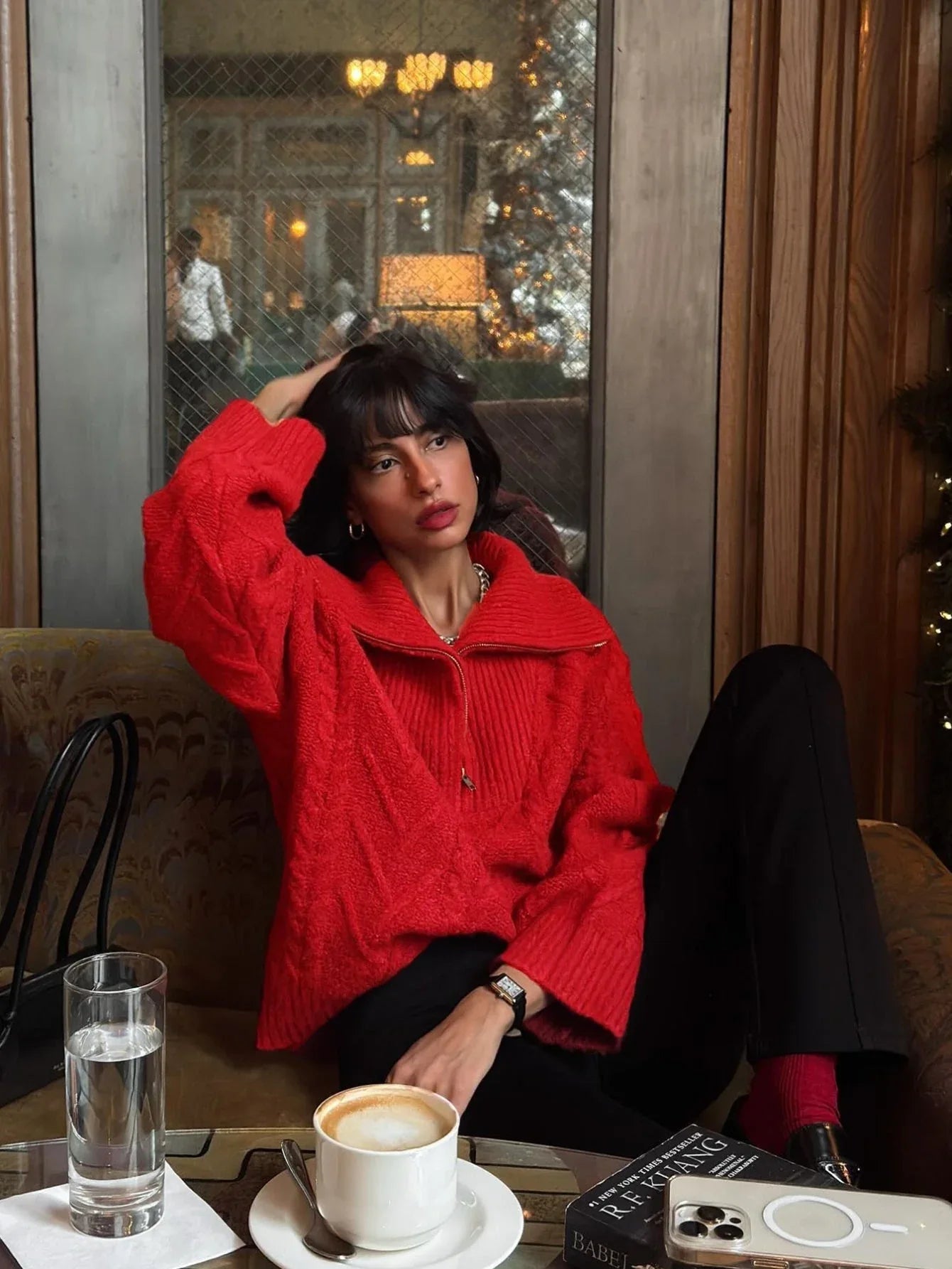 Woman sitting on a couch wearing a red textured knit quarter zip sweater and black pants, touching her hair.