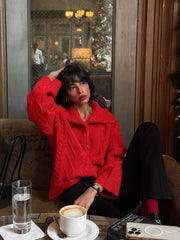 Woman sitting on a couch wearing a red textured knit quarter zip sweater and black pants, touching her hair.