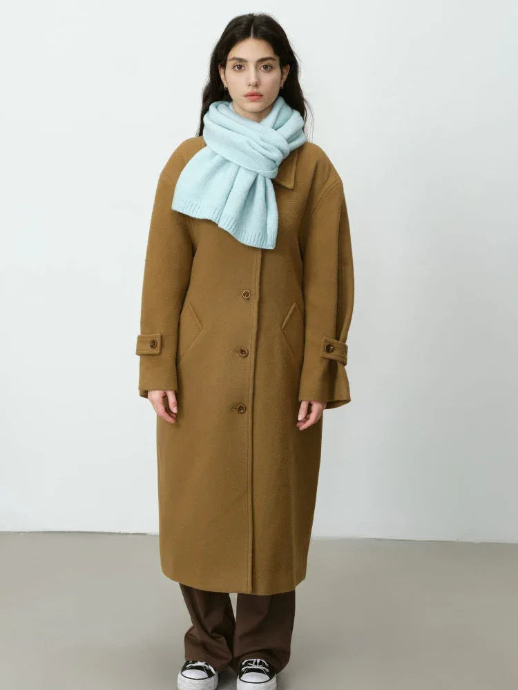 Full body shot of a woman wearing a long brown coat with a light blue scarf.
