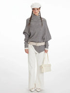 Woman wearing a grey asymmetrical knit poncho with a turtleneck, white pants, and a white belt, front view