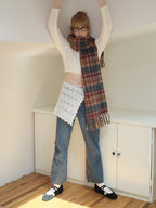 Model wearing a white cable knit cropped sweater, blue denim jeans, a plaid scarf, crochet overlay, and glasses, standing with arms raised.