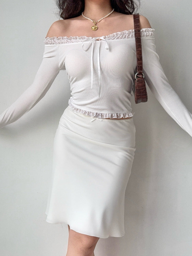 Woman wearing a white off-the-shoulder ribbed knit top with ruffled trim and tie front, paired with a white skirt and a brown handbag.