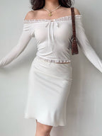 Woman wearing a white off-the-shoulder ribbed knit top with ruffled trim and tie front, paired with a white skirt and a brown handbag.