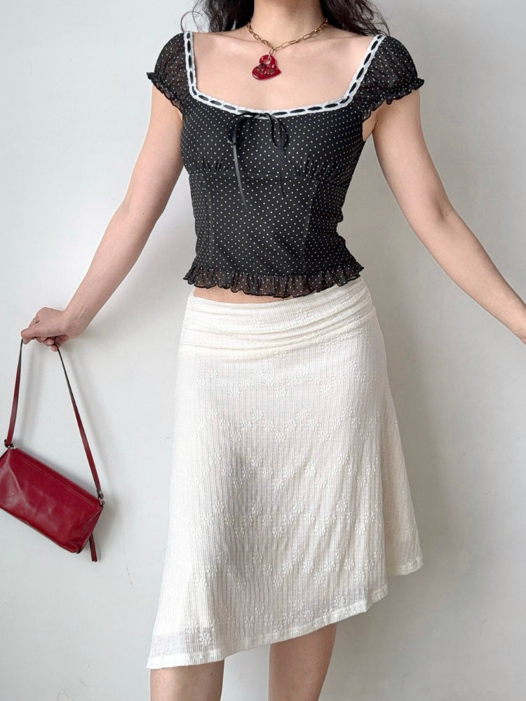 Full body shot of a woman wearing a black and white polka dot mesh top with a white textured skirt and a red handbag.