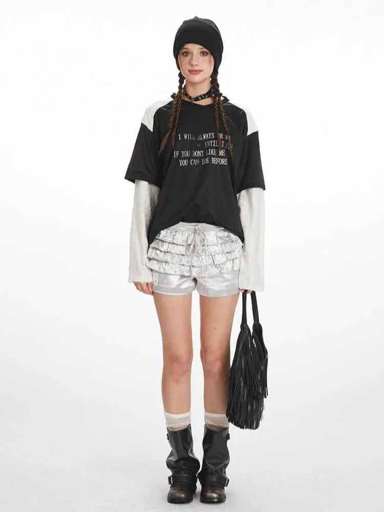 Full outfit view of a person wearing a layered black and white long-sleeved shirt with a graphic print, silver ruffled shorts, white socks, and black boots, holding a black fringed bag.