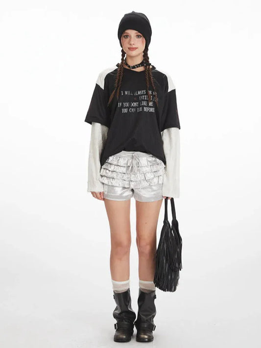 Full outfit view of a person wearing a layered black and white long-sleeved shirt with a graphic print, silver ruffled shorts, white socks, and black boots, holding a black fringed bag.