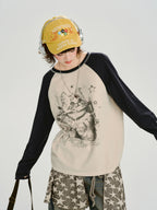 Front view of a model in a cream and navy raglan long-sleeve shirt with a cat graphic print and a yellow cap.