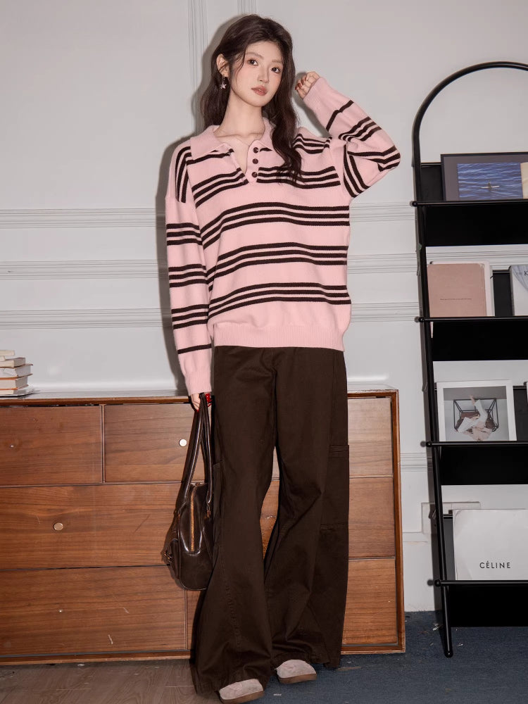 Full body shot of a woman wearing a pink and black striped knit polo sweater and brown pants, holding a handbag, standing in front of a dresser and a bookshelf.