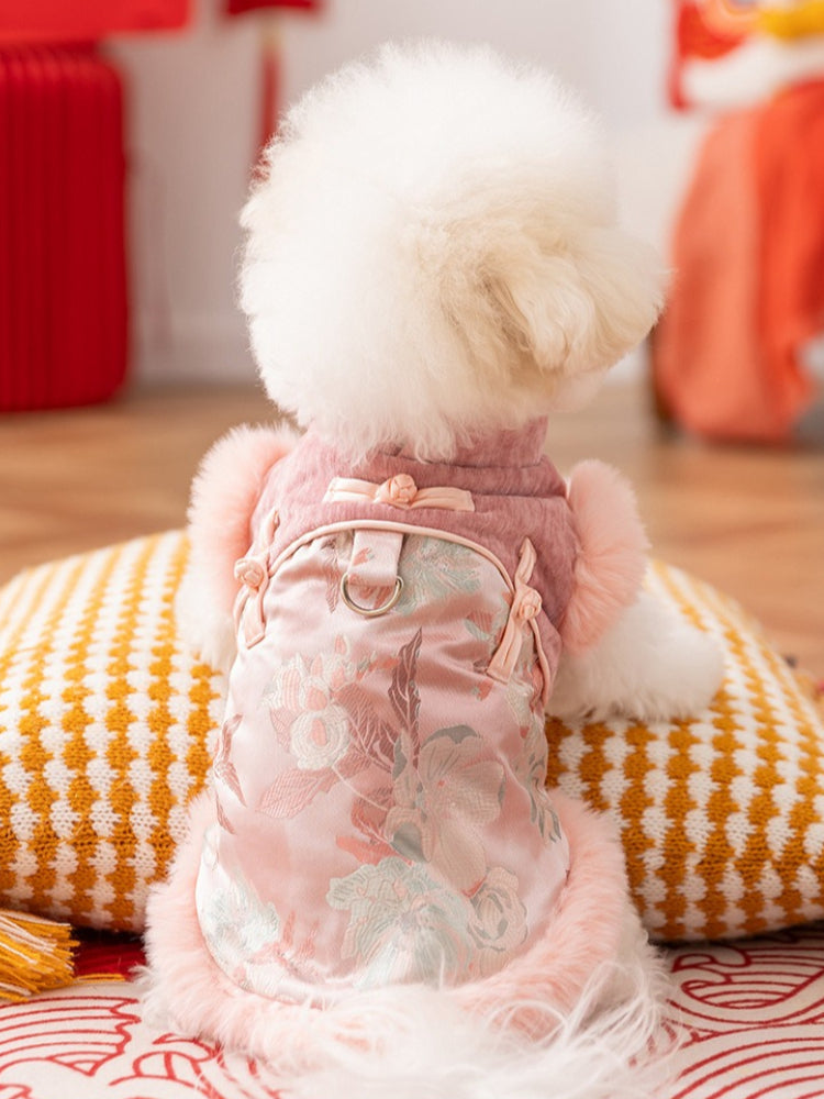 Rear view of a white Bichon Frise dog wearing a pink floral embroidered vest with faux fur trim, sitting on a cushion.