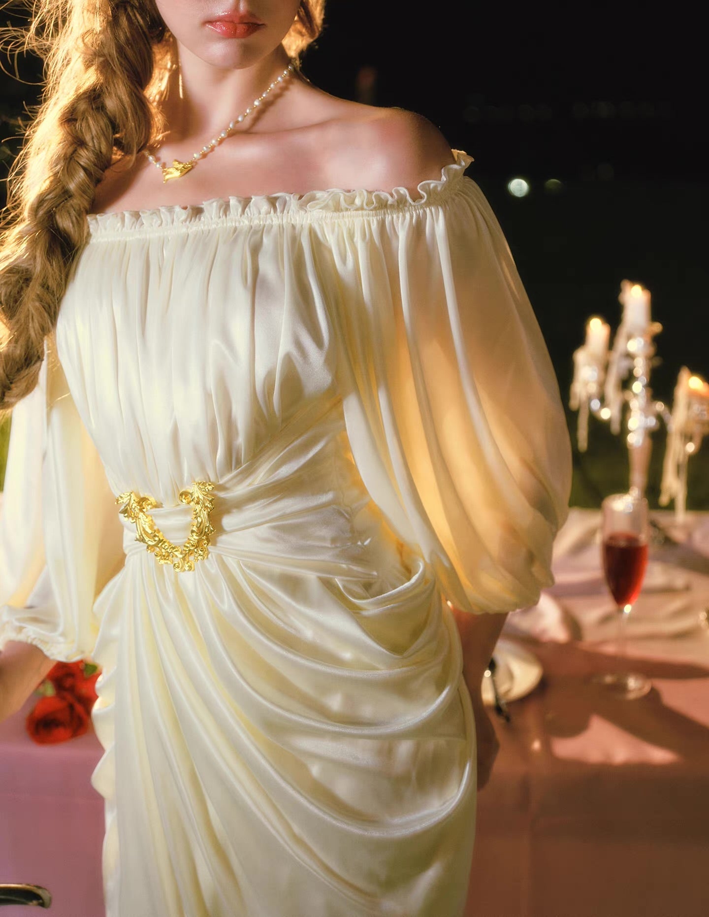 A woman poses with a sword while wearing a flowing cream satin gown with off-the-shoulder neckline and dramatic sleeves.