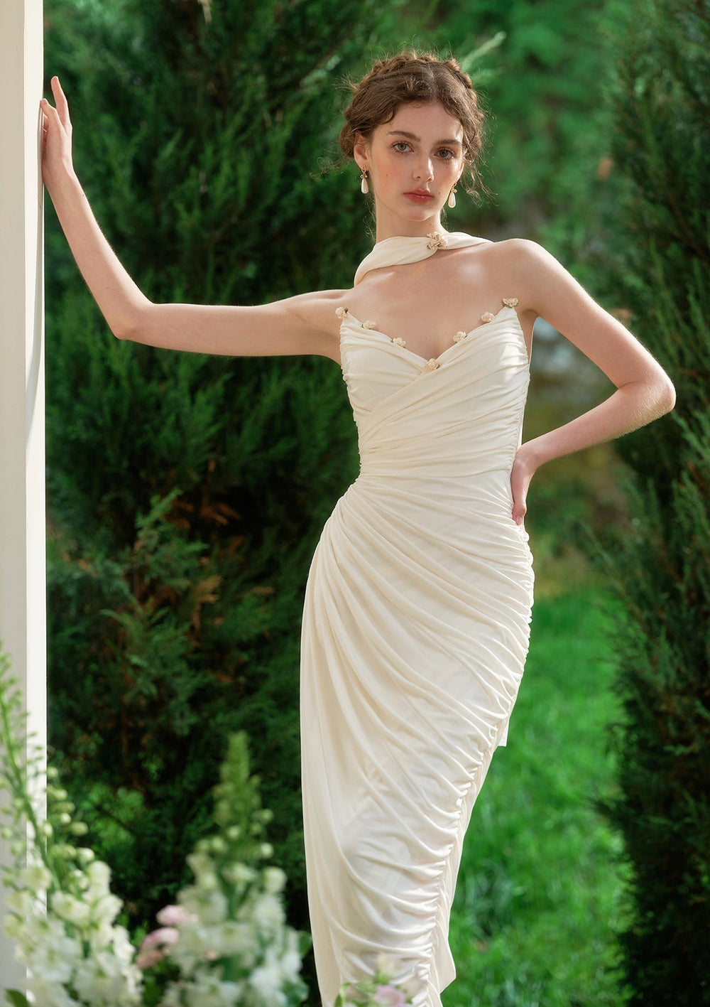 Woman in a cream satin mermaid gown with sweetheart neckline and floral embellishments, leaning against a white pillar outdoors.