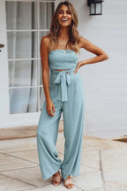 woman wearing a light blue strapless jumpsuit with wide-leg pants and a tie-waist detail, front view and smiling