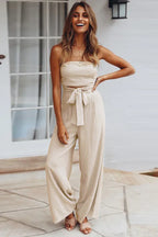 woman wearing a beige strapless jumpsuit with wide-leg pants and a tie-waist detail, standing outdoors and smiling