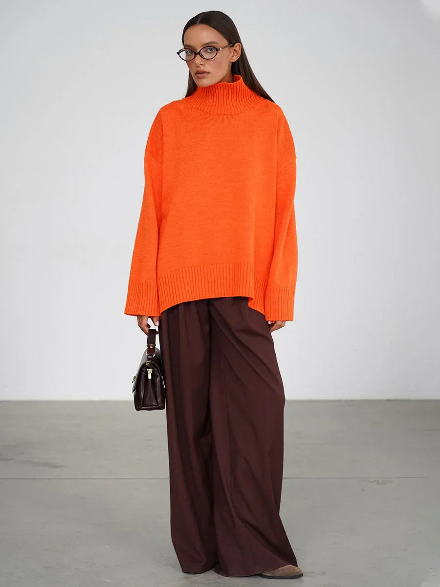 Woman wearing an oversized bright orange turtleneck sweater with wide-leg brown trousers and holding a dark handbag.