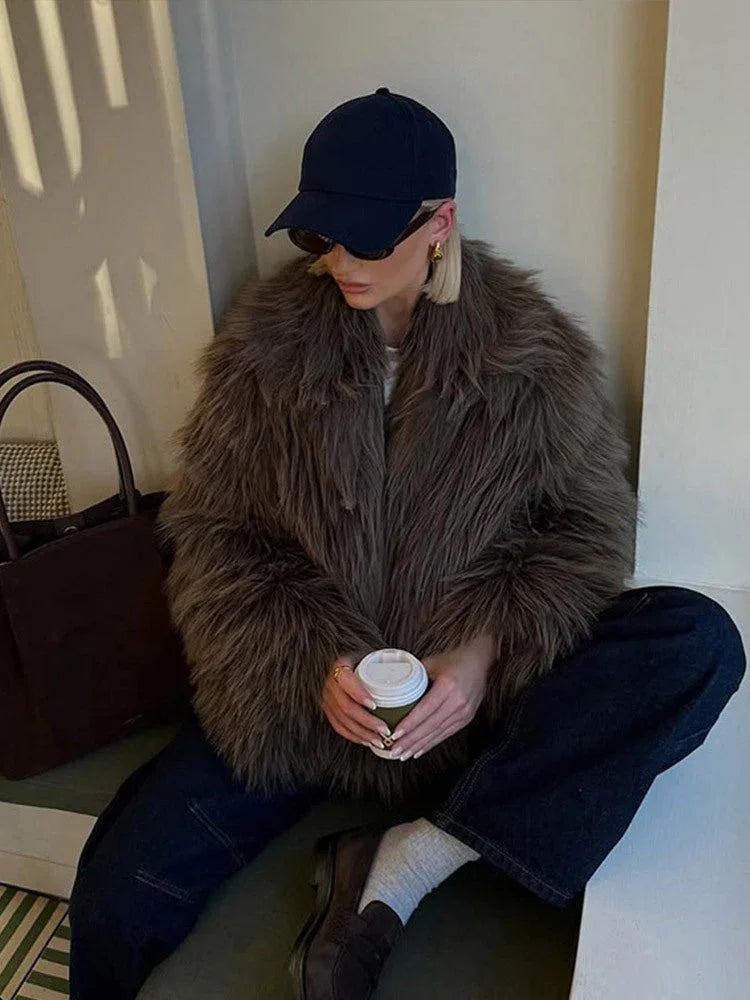 Woman sitting and holding a coffee cup, wearing a shaggy grey faux fur jacket, dark wash wide-leg jeans, and a navy baseball cap.