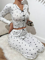 Woman wearing a white long-sleeve pajama set with a black heart print, holding a small metal cup, sitting on a white rug.