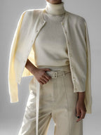 Woman wearing a cream-colored cashmere cardigan over a matching turtleneck and cream-colored trousers with a white belt.