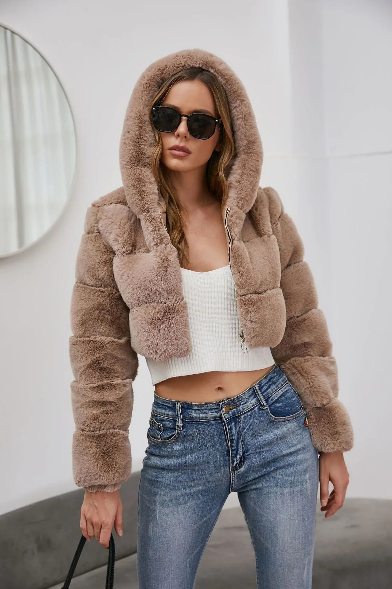 Woman wearing a cropped faux fur hooded jacket over a white camisole and blue jeans, front view.