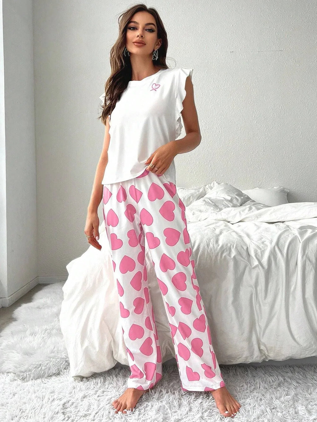 Woman standing next to a bed wearing white pajamas with pink heart print pants and a white top with a pink ribbon graphic.