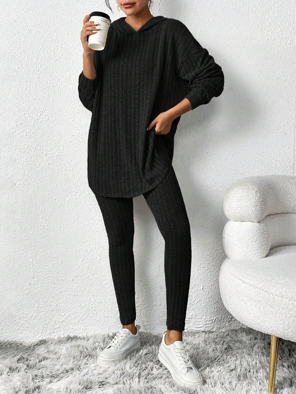 Woman in a black ribbed knit loungewear set, one hand in pocket, holding a coffee cup.