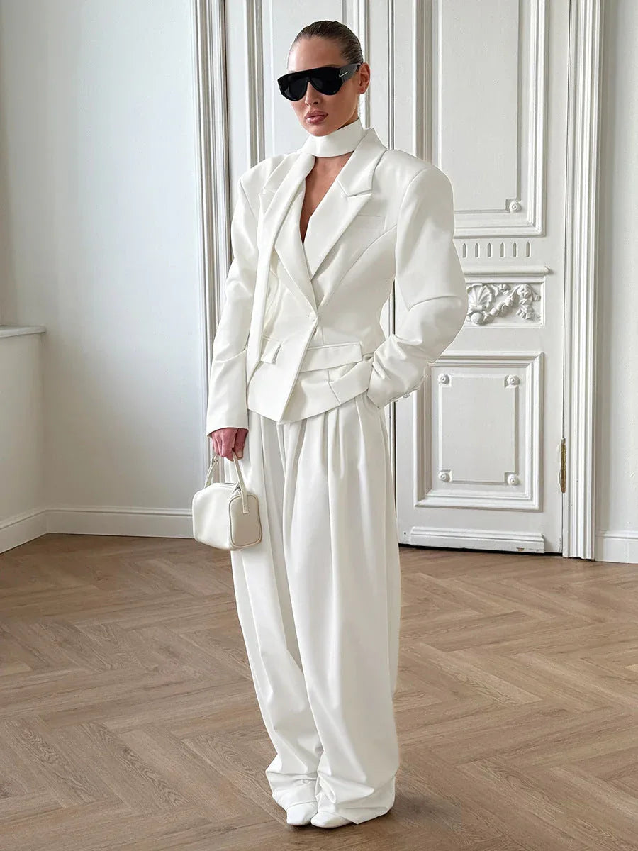 Full-body shot of a woman in a white oversized blazer and pleated trousers, styled with sunglasses and a handbag.