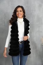 Woman wearing a black faux fur vest over a white turtleneck and blue jeans, smiling.