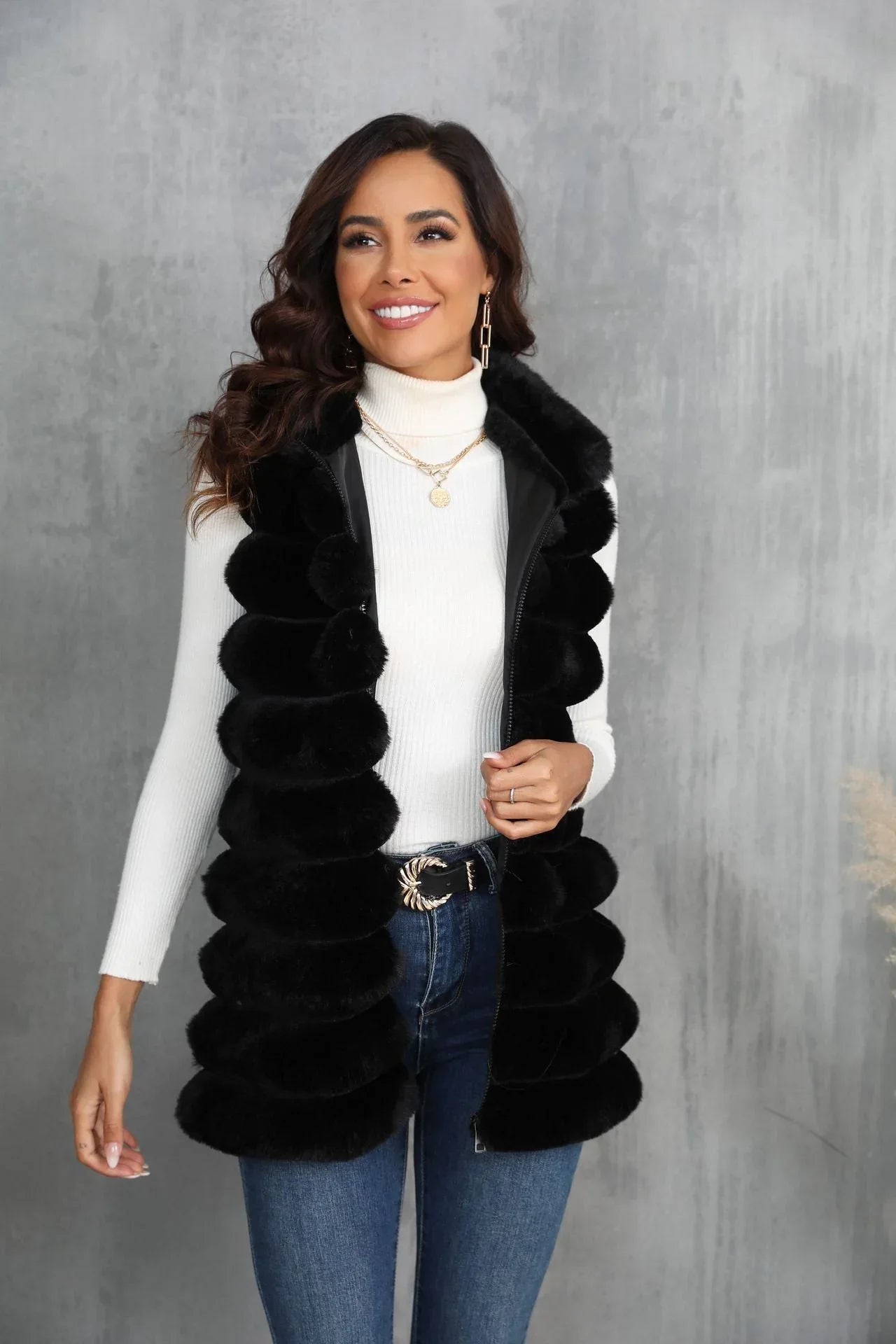 Woman wearing a black faux fur vest over a white turtleneck and blue jeans, smiling.