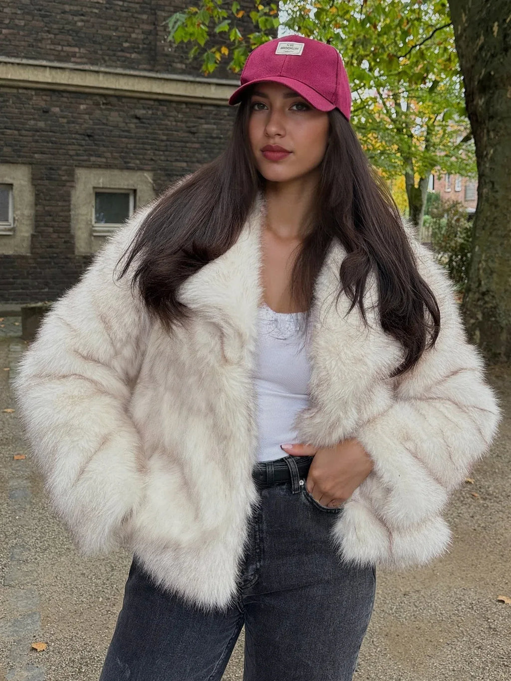 Woman in a cream faux fur jacket and pink baseball cap, posing outdoors.