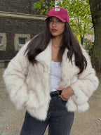 Woman in a cream faux fur jacket and pink baseball cap, posing outdoors.