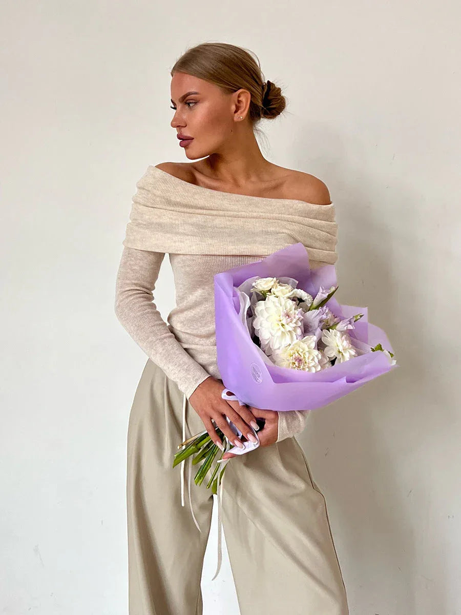 Woman wearing a cream off-the-shoulder ribbed knit sweater with long sleeves and beige wide-leg pants, holding a bouquet of white and purple flowers.