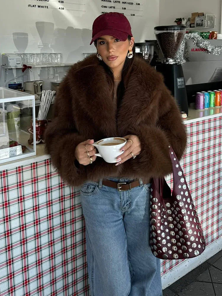 woman wearing a brown faux fur coat with a baseball cap and jeans, holding a coffee cup in a cafe