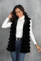 Woman wearing a black faux fur vest over a white turtleneck and blue jeans, touching her hair and smiling.
