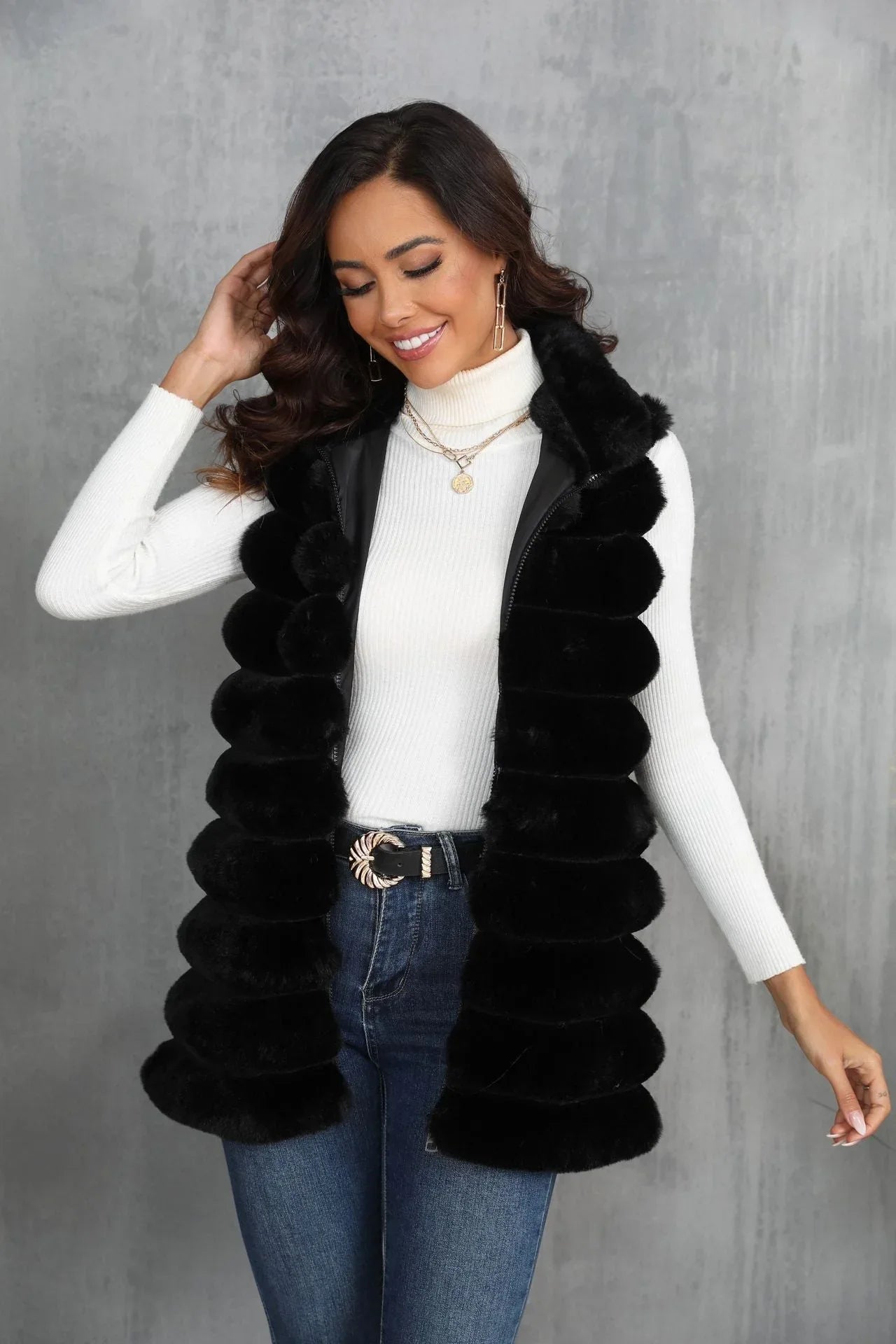 Woman wearing a black faux fur vest over a white turtleneck and blue jeans, touching her hair and smiling.