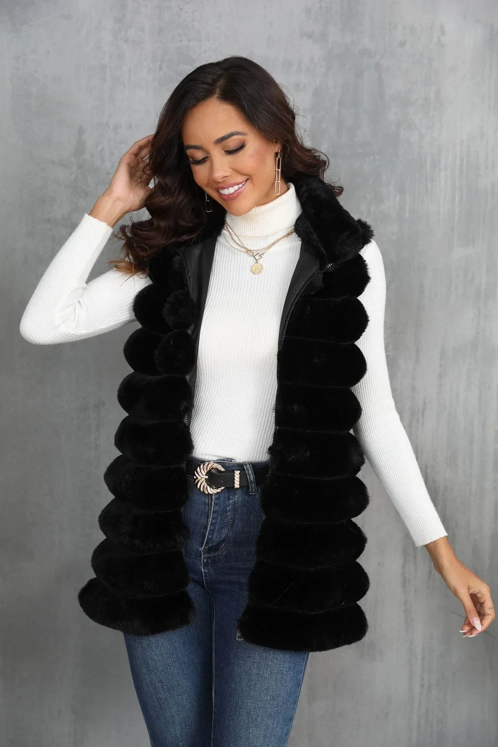 Woman wearing a black faux fur vest over a white turtleneck and blue jeans, touching her hair and smiling.