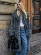 Woman wearing a long grey faux fur coat and blue jeans, leaning against a stone wall.