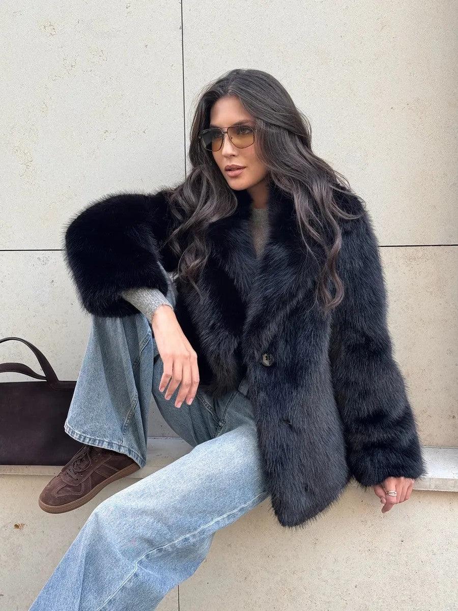Woman wearing a black faux fur jacket over a grey top and jeans, sitting down.