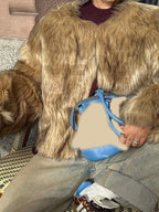 Close-up of a woman's outfit featuring a tan faux fur jacket, burgundy sweater, and a blue and tan handbag.