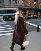 Woman walking away from the camera, wearing a long brown faux fur coat and carrying a white quilted handbag, with a large white bow in her hair.
