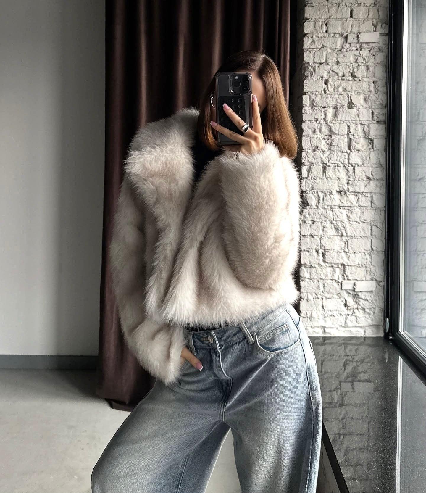 Side view of a woman in a cream faux fur cropped jacket and light wash wide-leg jeans, holding a phone to take a mirror selfie.