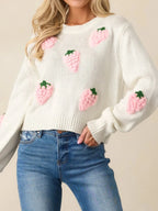 Front view of a white cropped knit sweater adorned with pink strawberry appliques, paired with blue denim jeans.