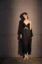 Woman wearing a black satin slip dress with lace trim and a matching robe, standing against a grey wall.