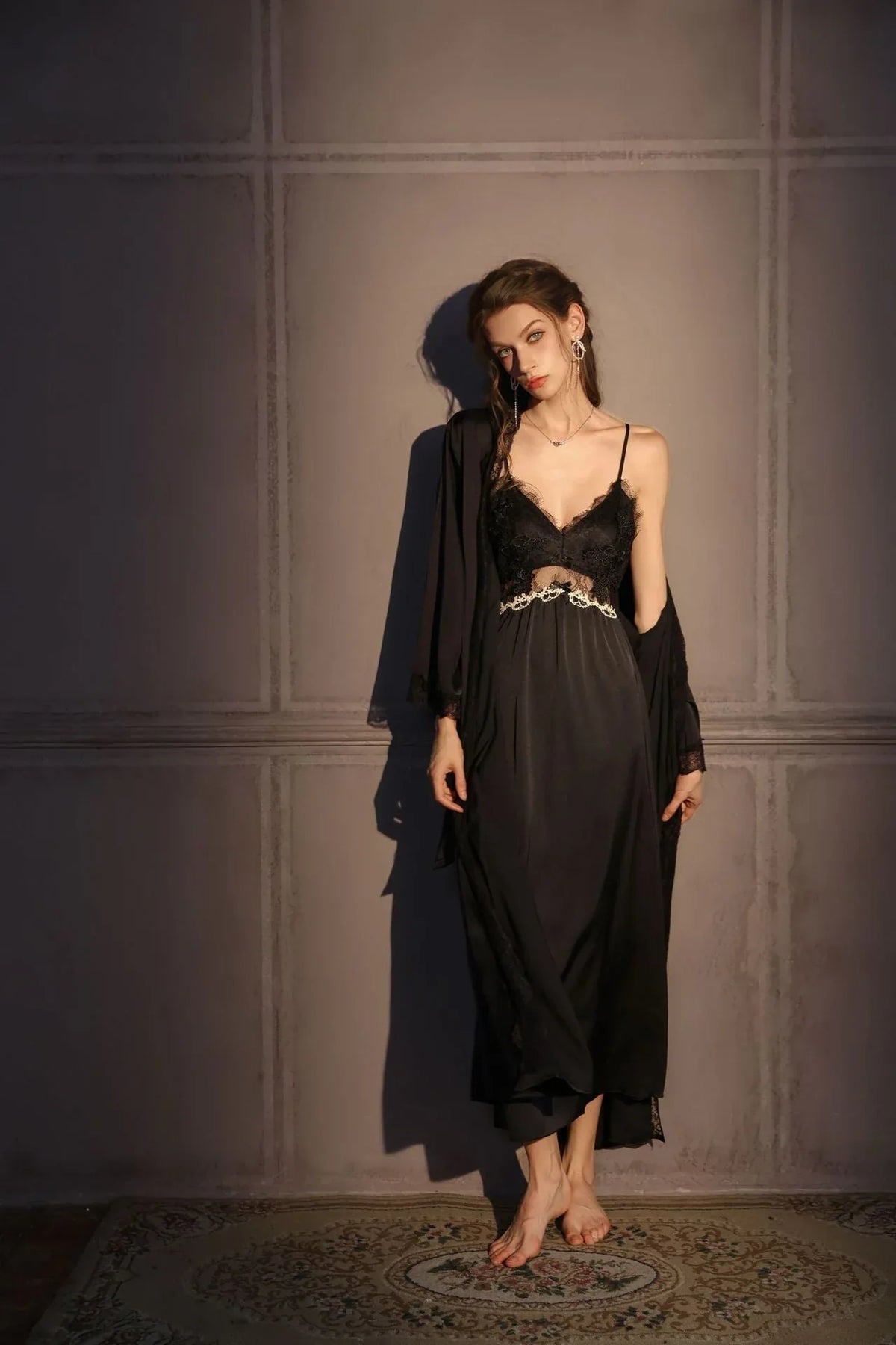 Woman wearing a black satin slip dress with lace trim and a matching robe, standing against a grey wall.