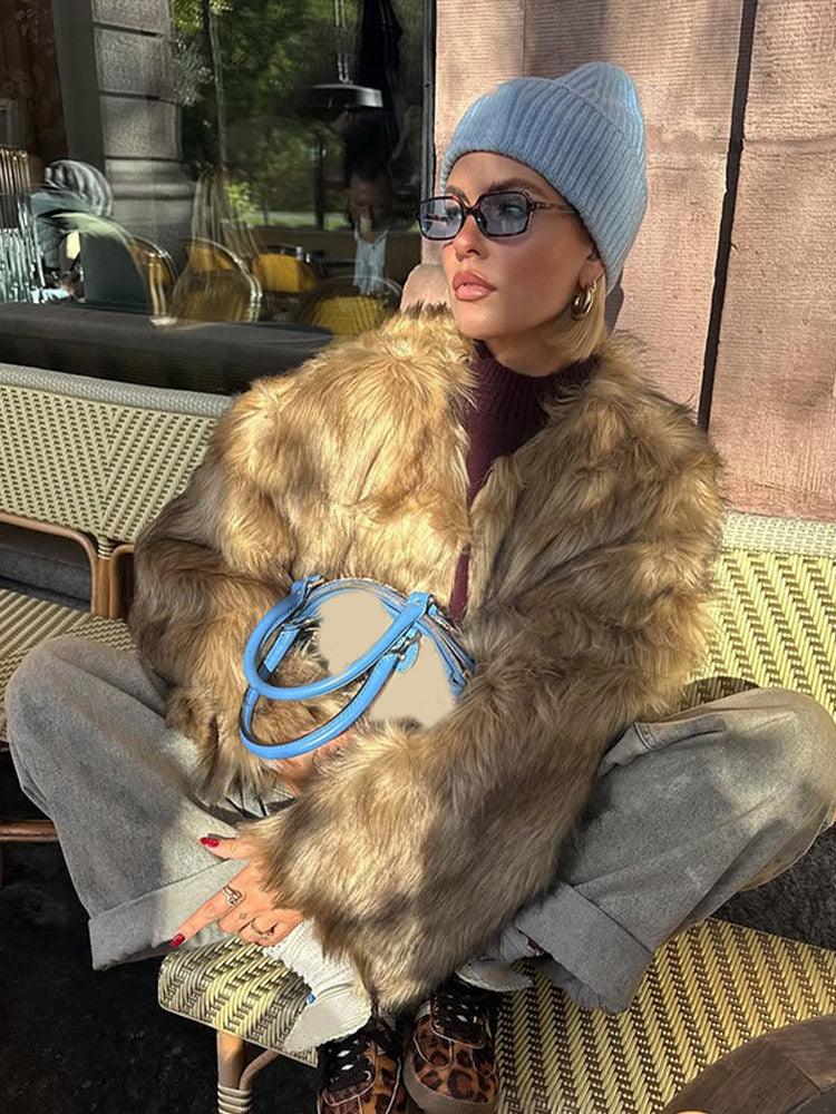 Woman sitting outdoors wearing a tan faux fur jacket, blue beanie, and grey pants, holding a blue and tan bag.