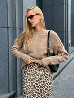 Woman adjusting her leopard print skirt while wearing a tan cable knit sweater and sunglasses.