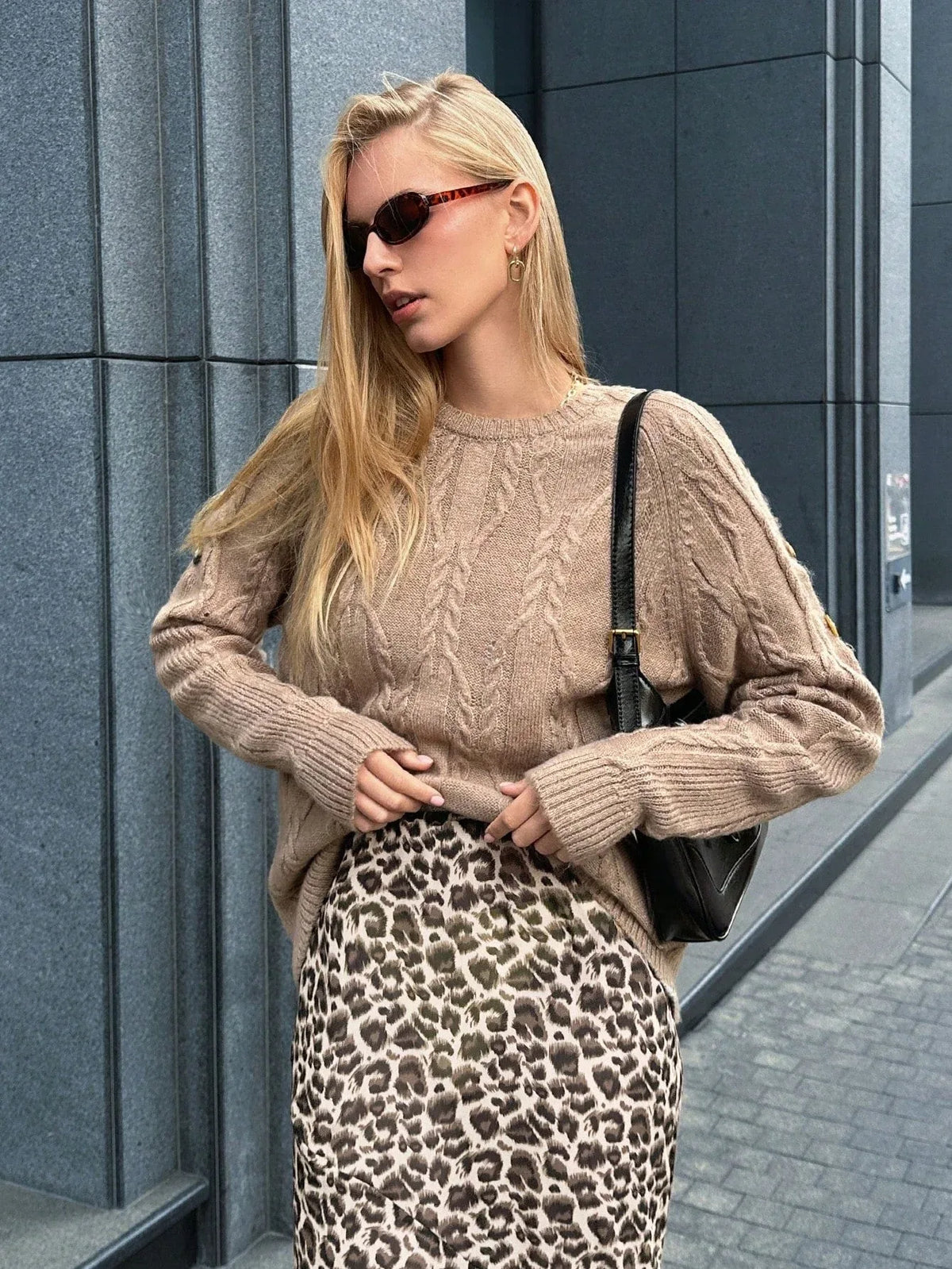 Woman adjusting her leopard print skirt while wearing a tan cable knit sweater and sunglasses.