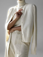Woman wearing a cream-colored cashmere cardigan over a matching turtleneck and cream-colored trousers, arms crossed.