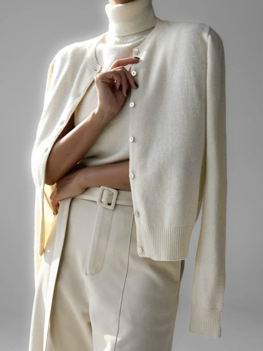Woman wearing a cream-colored cashmere cardigan over a matching turtleneck and cream-colored trousers, arms crossed.