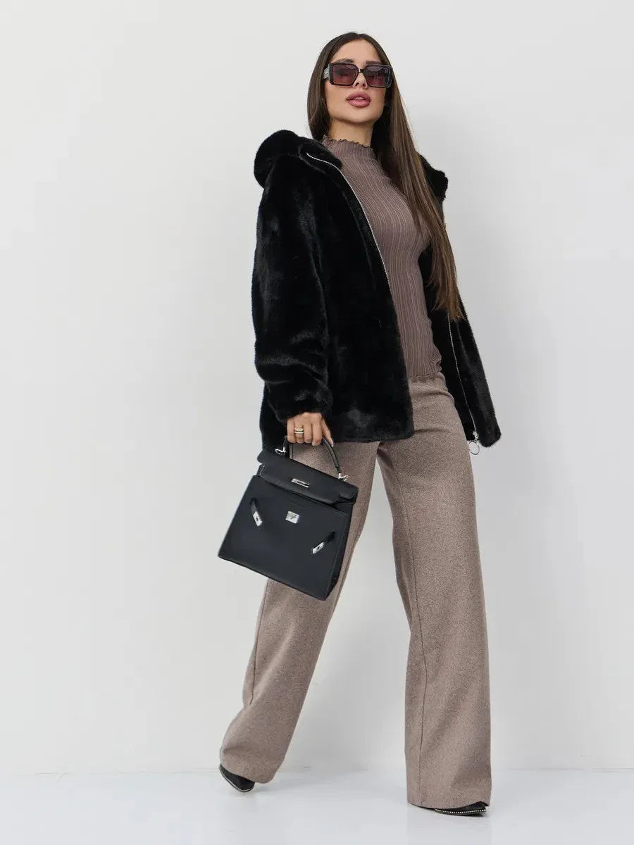 Full-body shot of a woman wearing a black hooded faux fur jacket, brown ribbed turtleneck top, and brown wide-leg trousers, holding a black handbag and wearing sunglasses.