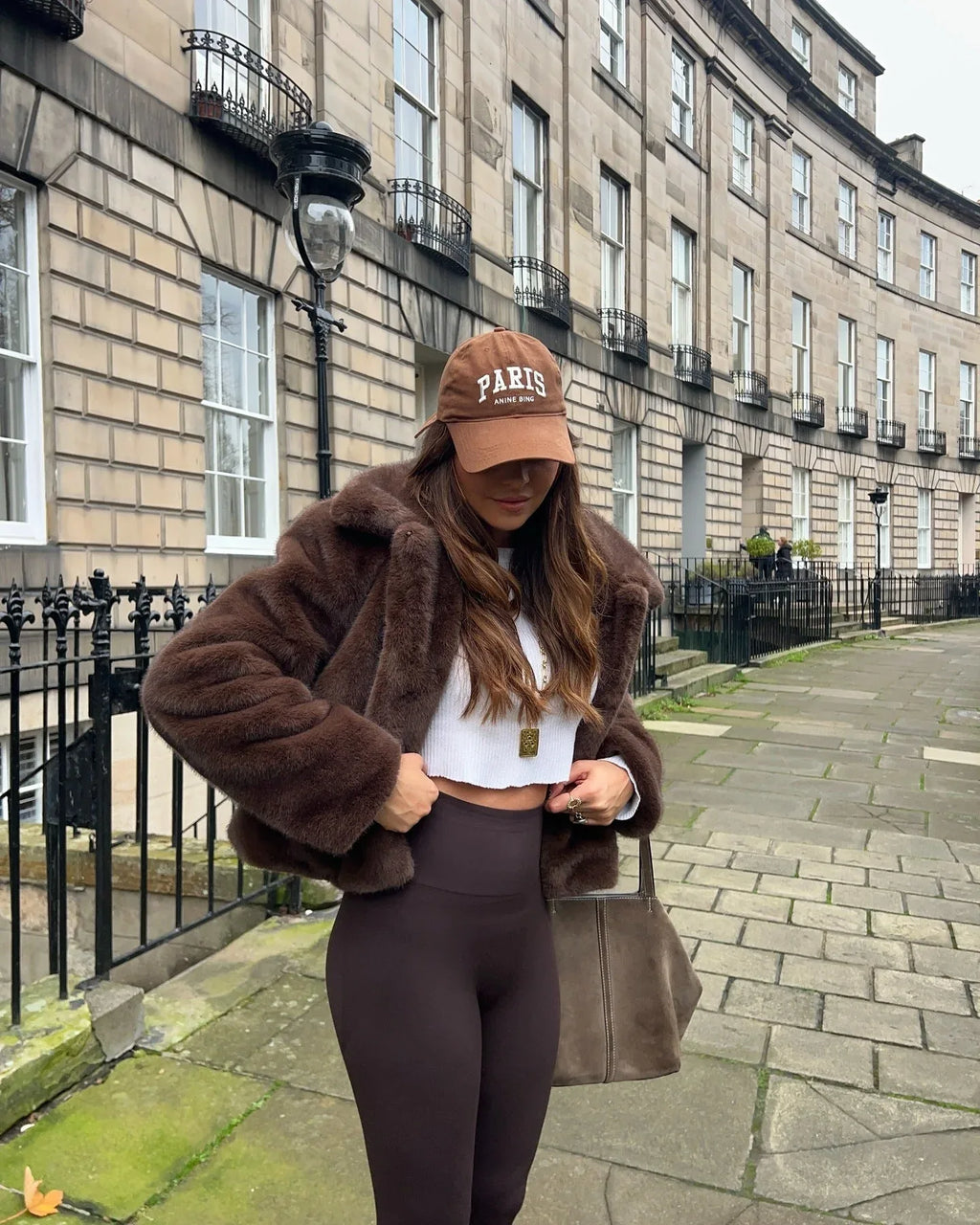 Woman wearing a brown faux fur cropped jacket over a white crop top and brown leggings, holding her hands to the jacket.